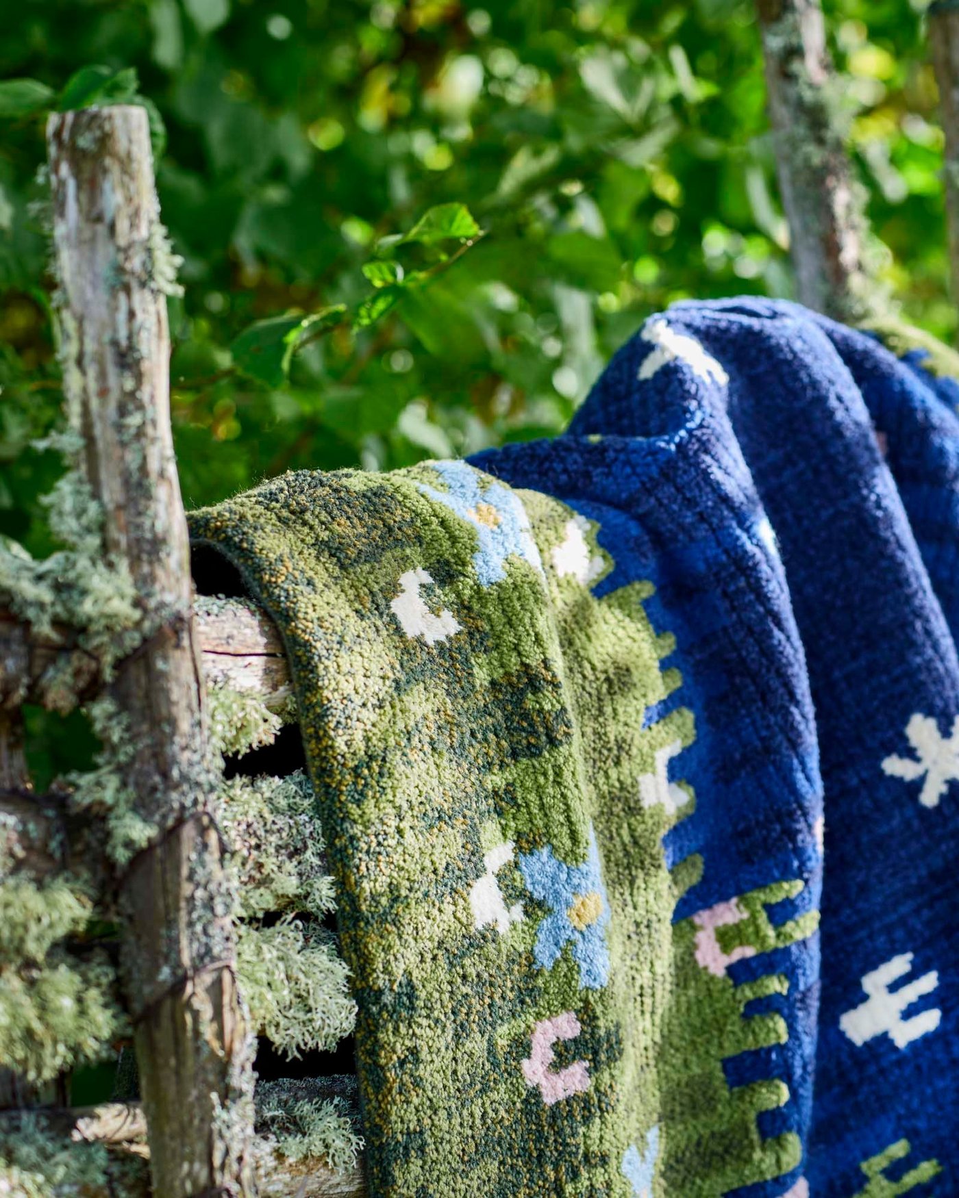 A cropped shot of a GRODDSVINGEL rug draped over a moss-covered wood fence, with trees in the background.