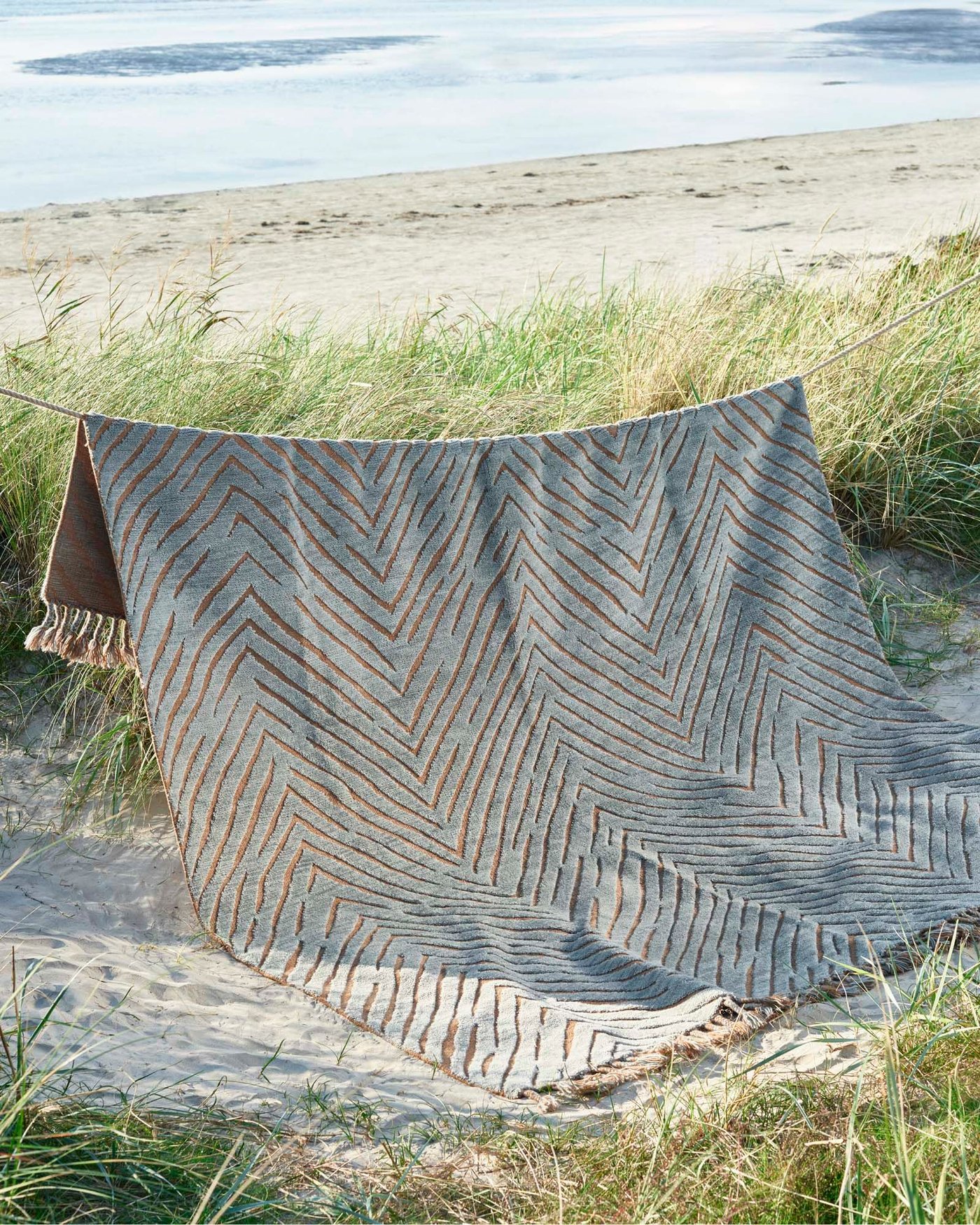 A KLAPPERSTARR rug hangs over a rope line, among the grass and sand on a beach, with the sea in the background.