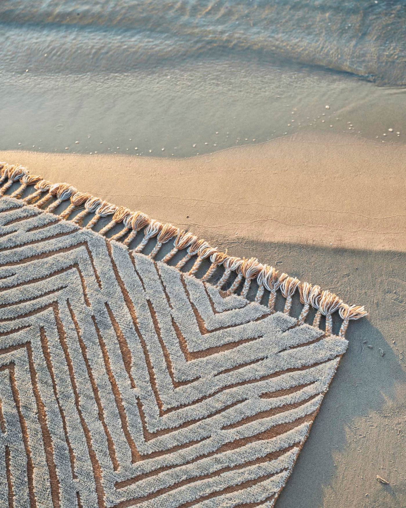 The corner of a KLAPPERSTARR rugs with tassles, laid out on a sandy surface with shadows.