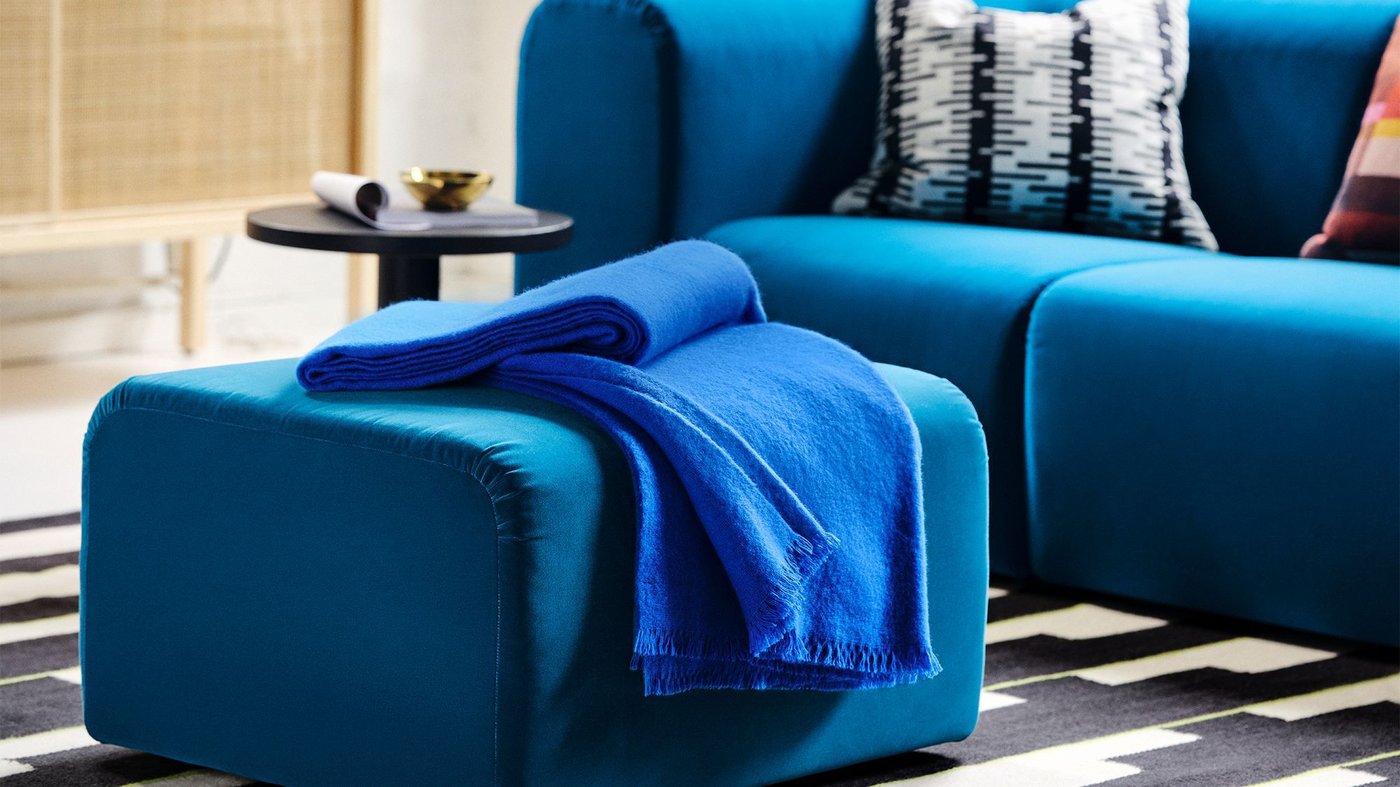 A blue velvet STOCKHOLM sofa and footstool in a white living room on a black and white rug.