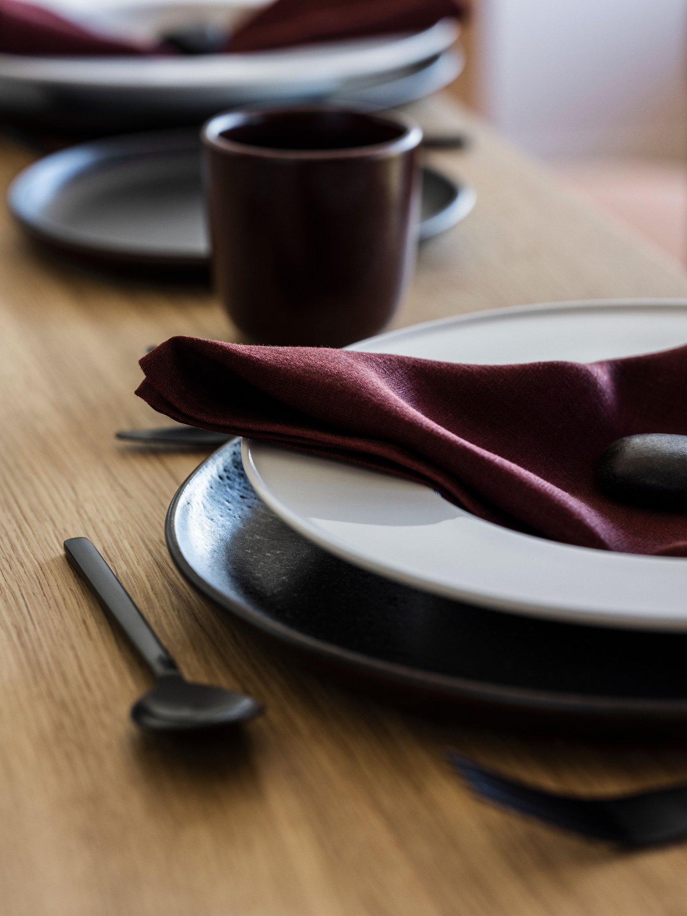 Close-up of stone-glazed diningware on wooden table, with burgundy linen cloth and utensils to the side.