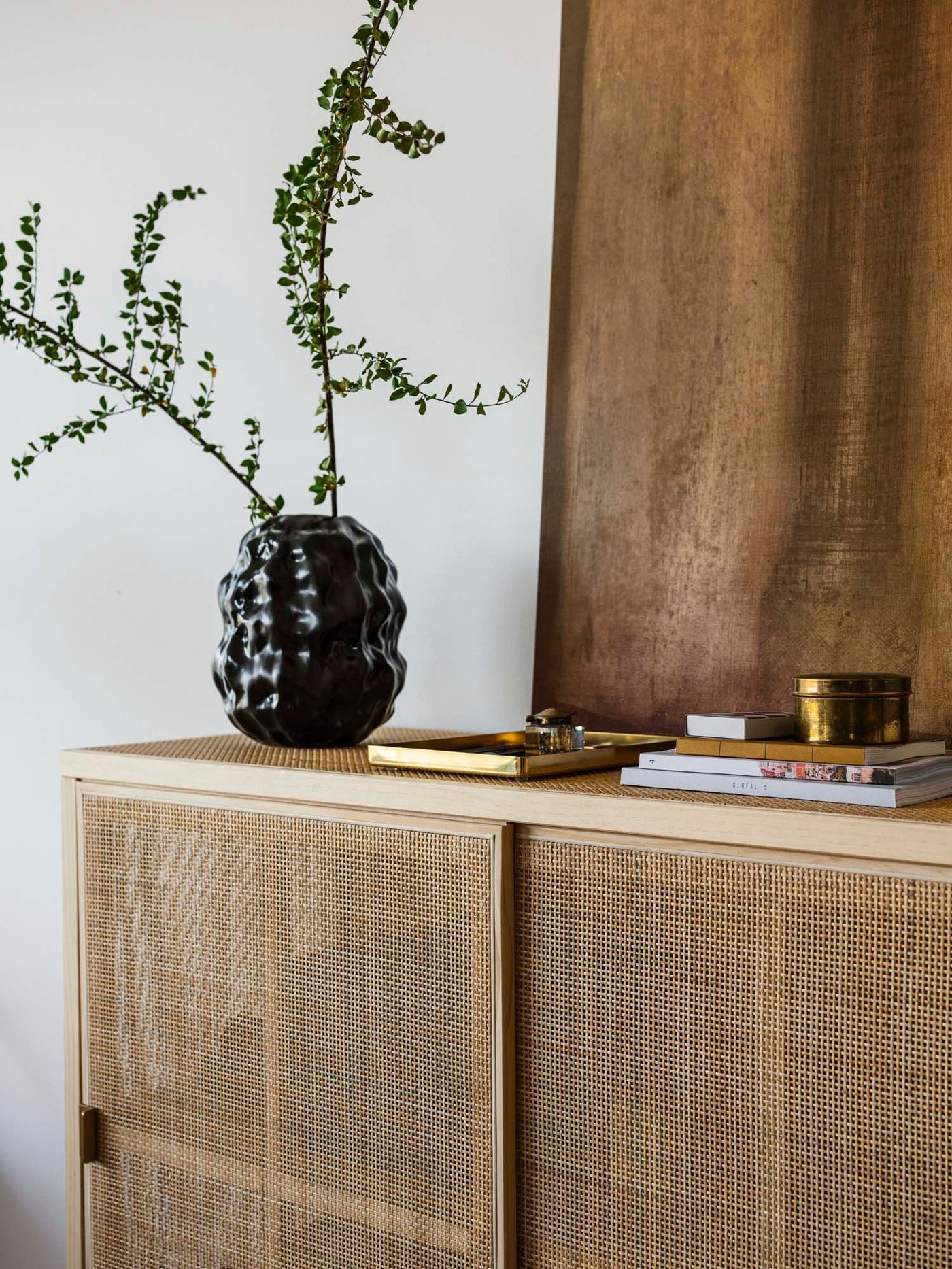 A black organic shaped vase with green branches sits on a rattan storage unit.