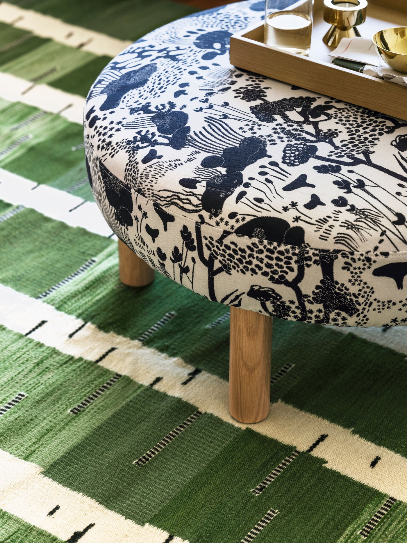 Mushroom-patterned pouffe with wooden legs on a green birch-patterned rug, topped with a wooden tray holding gold bowls, glass, and a gold candle holder.