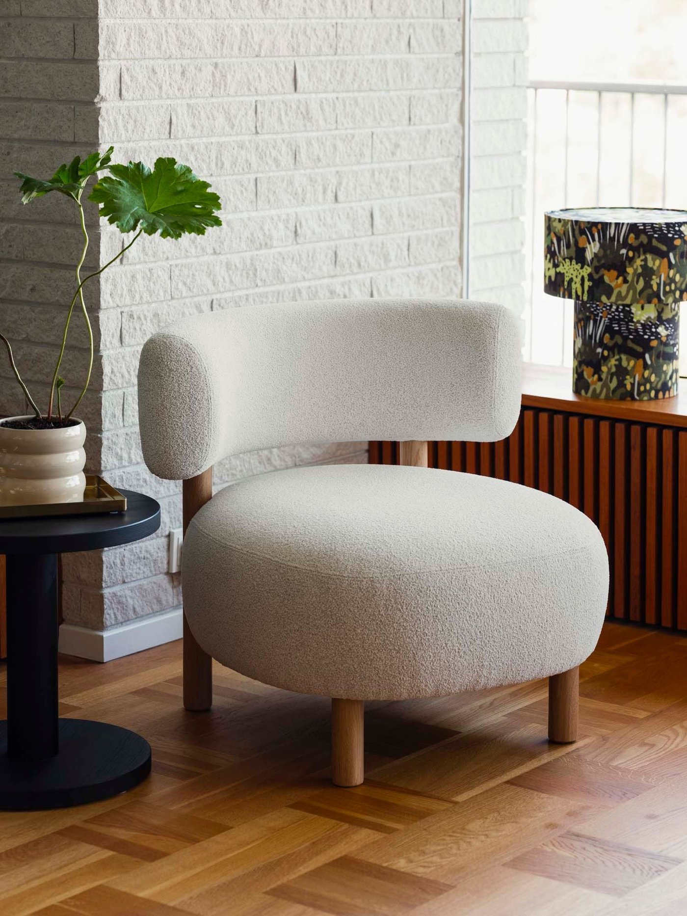 A round, upholstered chair from the collection stands on a wood floor next to a plant and floral table lamp.