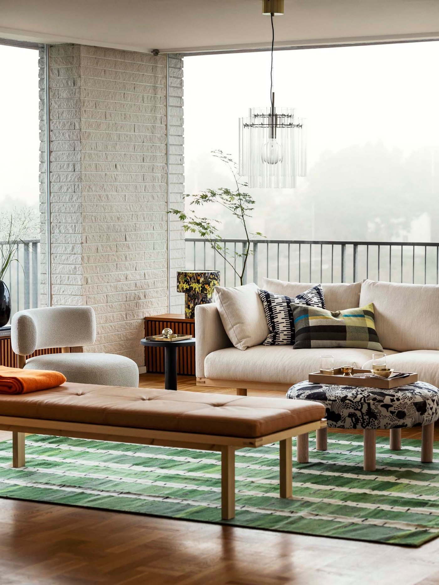 A modern living room with an armchair and sofa, a pouffe in a black and white pattern, a leather bench, on a green patterned rug.