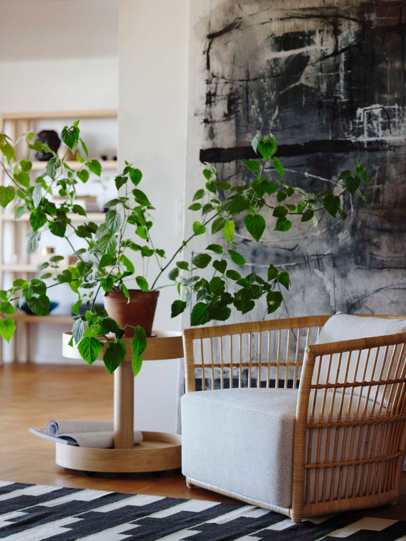 A armchair in rattan is standing on a black and white carpet, with green plant and coffee table next to it with a large painting in the back.