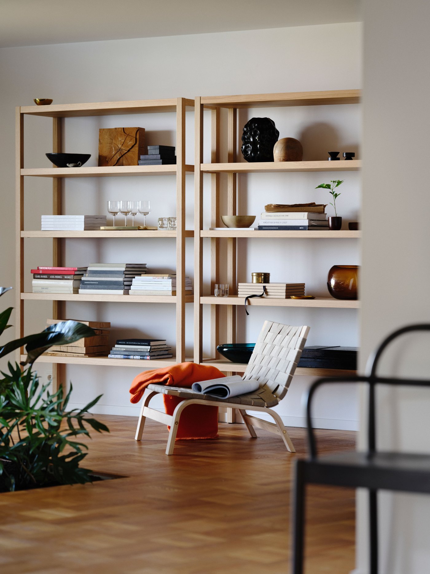 Two wide oak shelves with books, plants, vases, and bowls, saddle weave chair with orange throw in front.