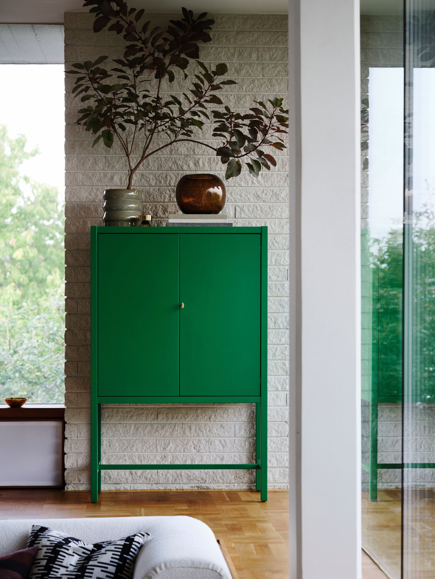 A veneer cabinet on two legs in green, with vases and flowers on top, standing in the back of a room, with parts of a window seen to the side.