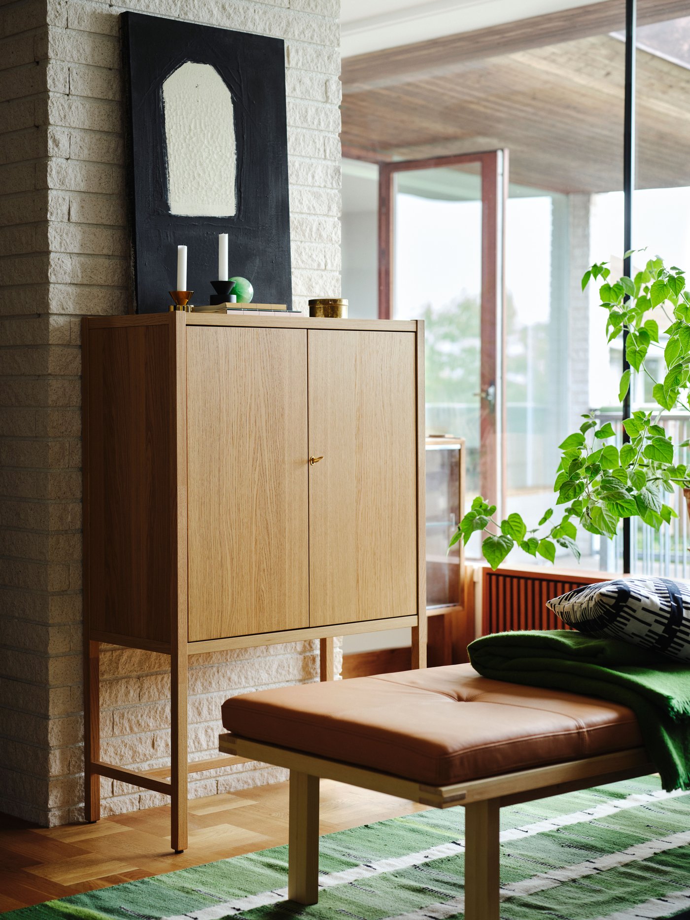 A wooden oak cabinet in the back of a room, with a daybed in pine with a cushion of seglora leather, standing on a green birch pattern rug.