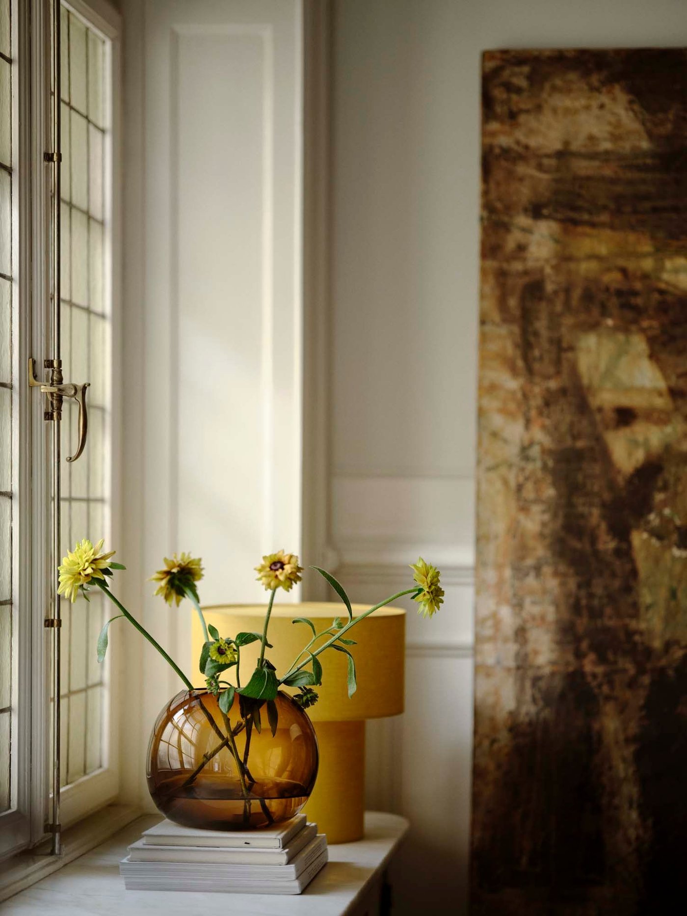 A yellow STOCKHOLM 2025 table lamp behind a brown STOCKHOLM 2025 vase with yellow flowers, beside a window.