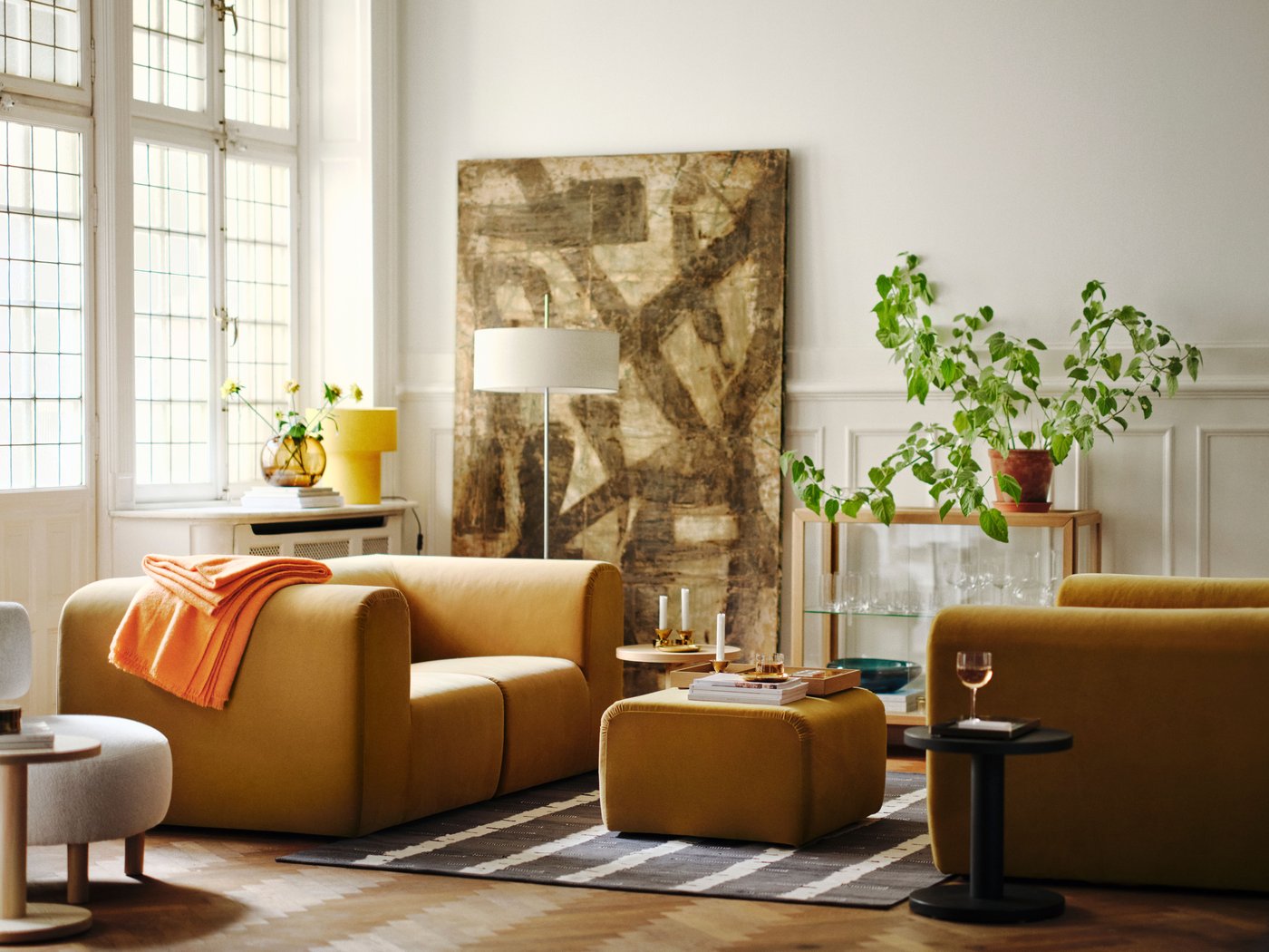 Two light brown sofas with matching pouffe and glass cabinet, set in a room with green plants, a large painting, and decorative vases.
