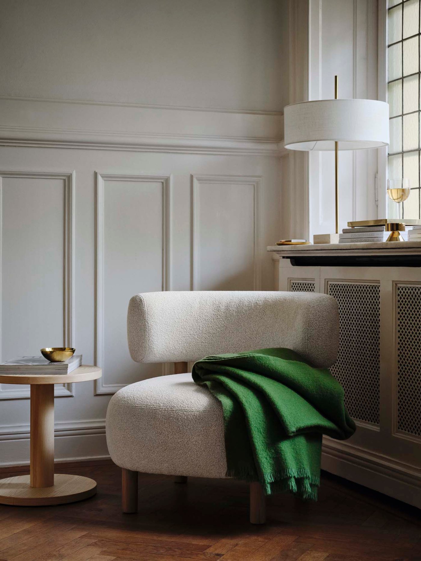 Light bouclé armchair with green wool blanket draped over, side table next to it, and a table lamp by the window in the background.