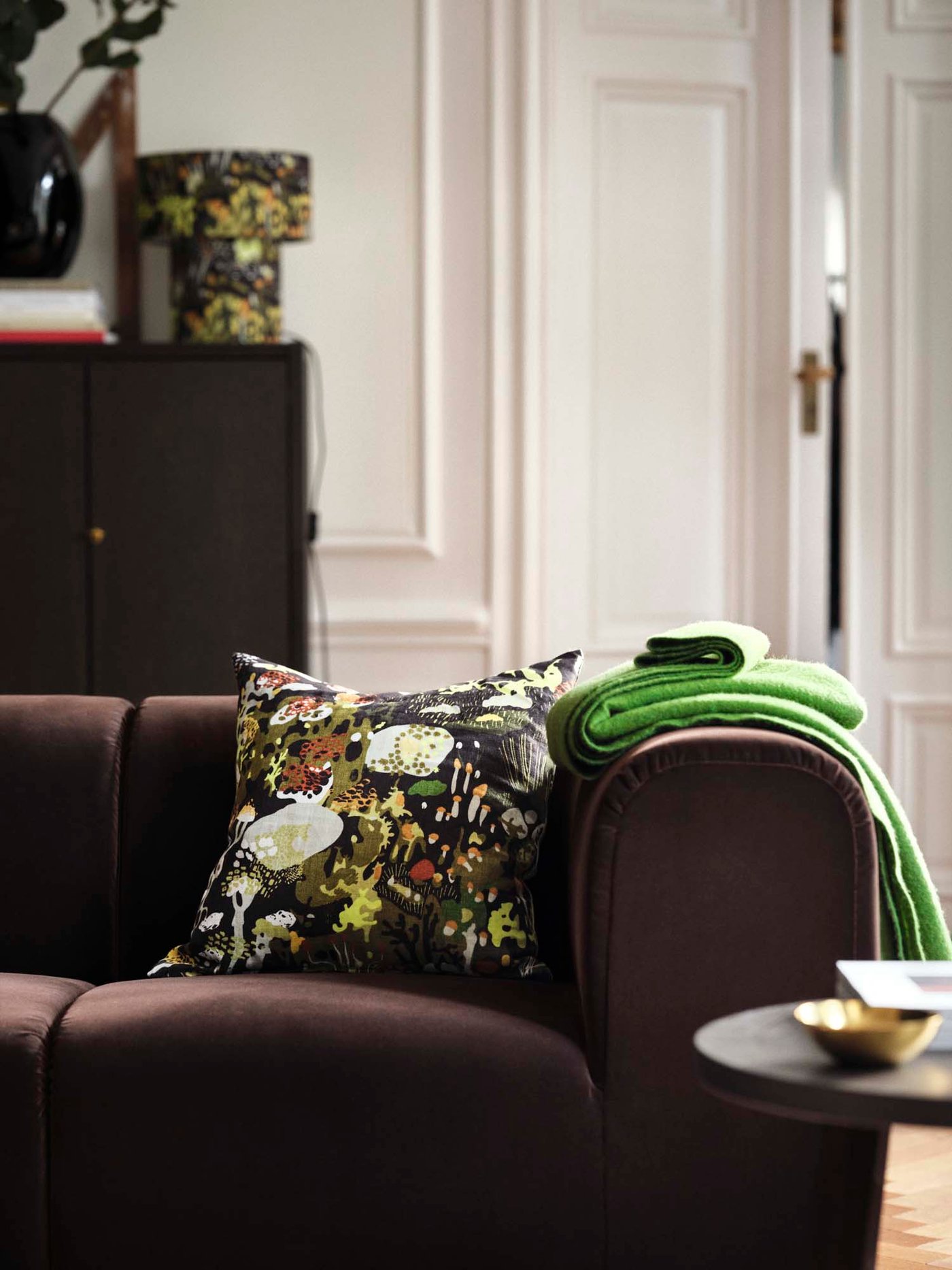Side view of dark brown sofa with green throw on armrest and forest-patterned pillow. Matching lamp in the back on a dark oak cabinet.