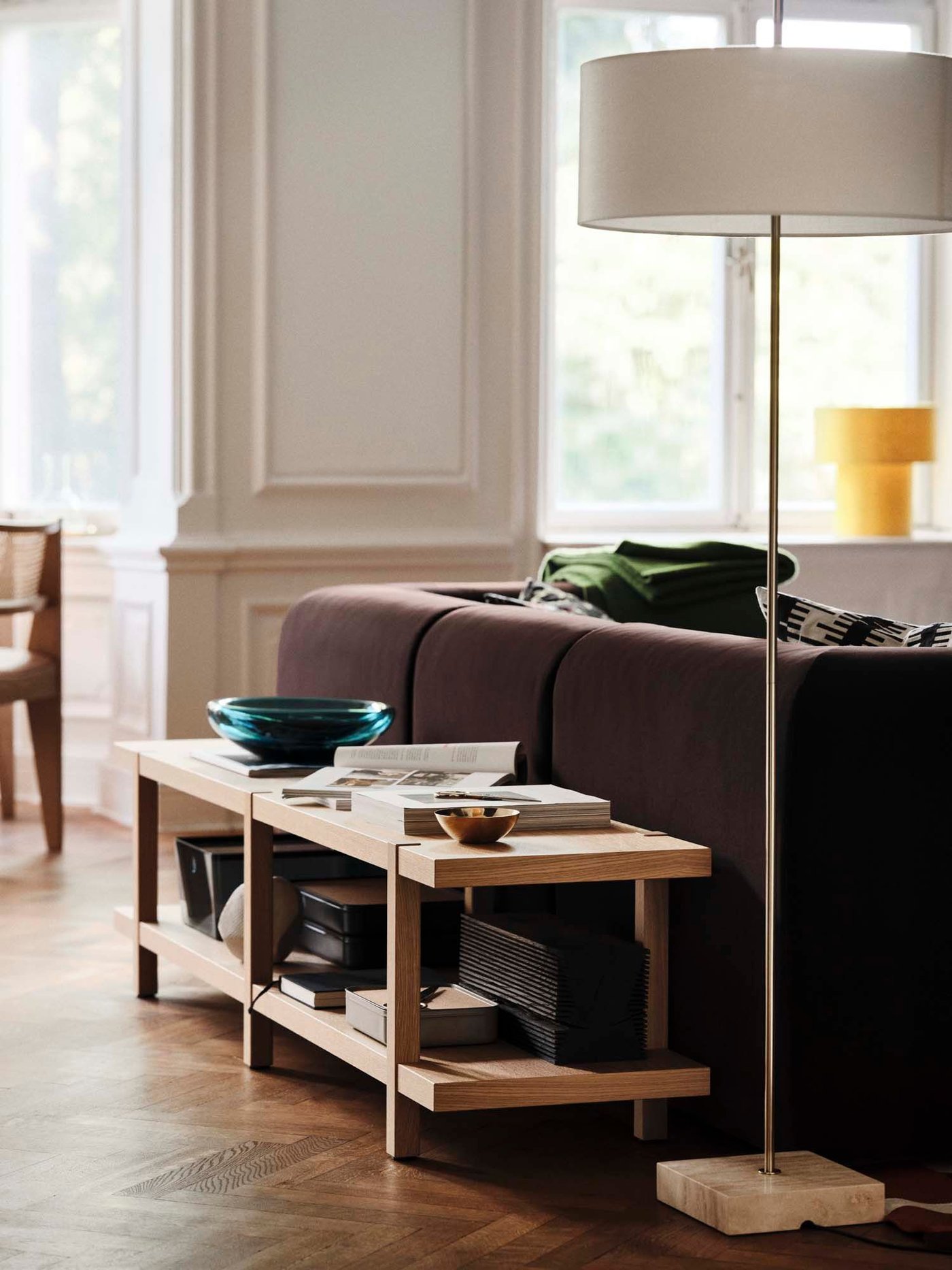 A shelving unit with glassware stands next to a tall lamp and behind a brown velvet sofa in a room.