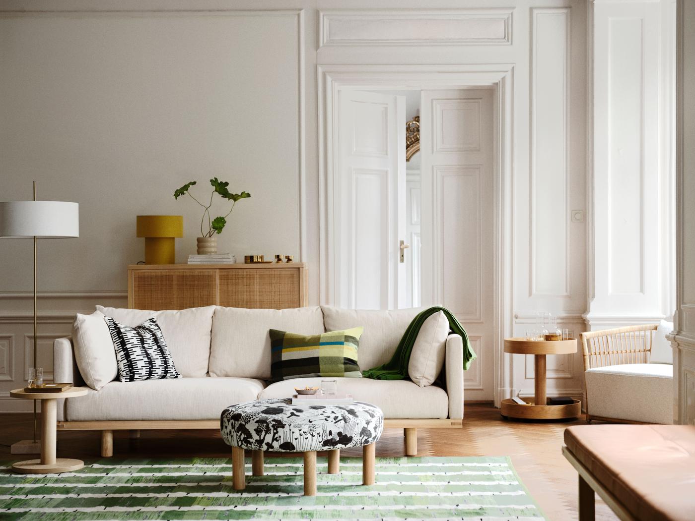 White sofa in beige wool on a green flatweave rug, with a pouffe, lamp, rattan cabinet, armchair in the corner, and daybed in the front.