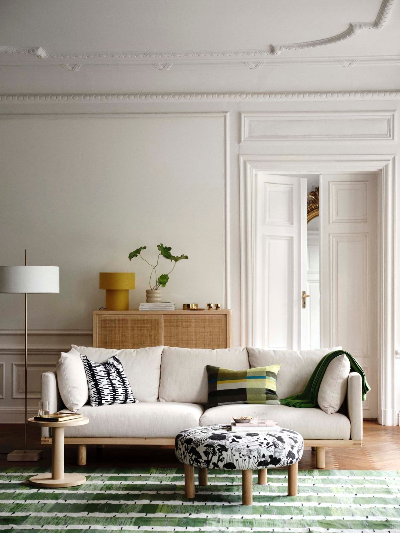 A white living room with light wood and light coloured furniture on a green patterned rug.
