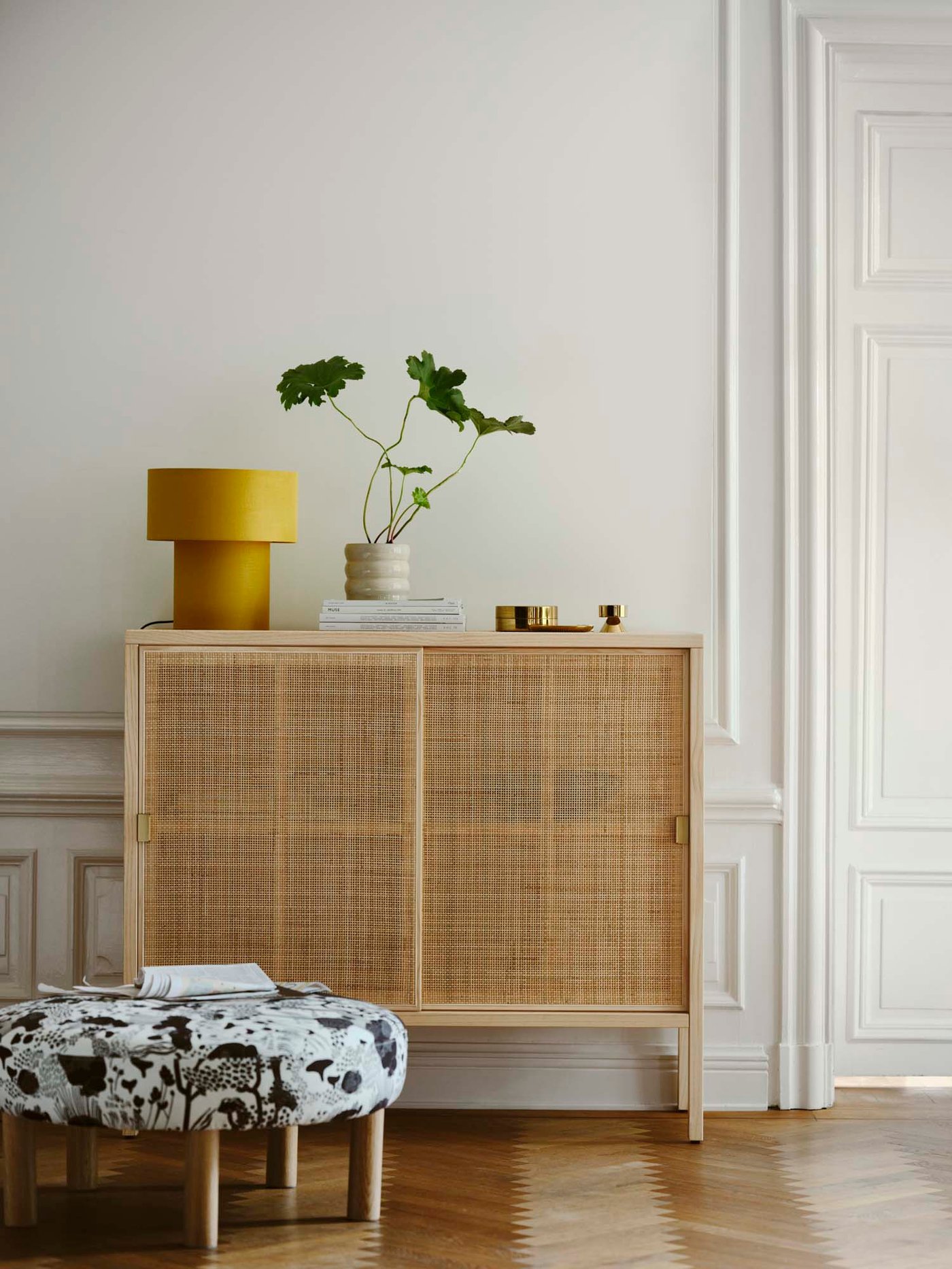 A rattan cabinet with a yellow table lamp on top, and a mushroom-pattern pouffe in front. 