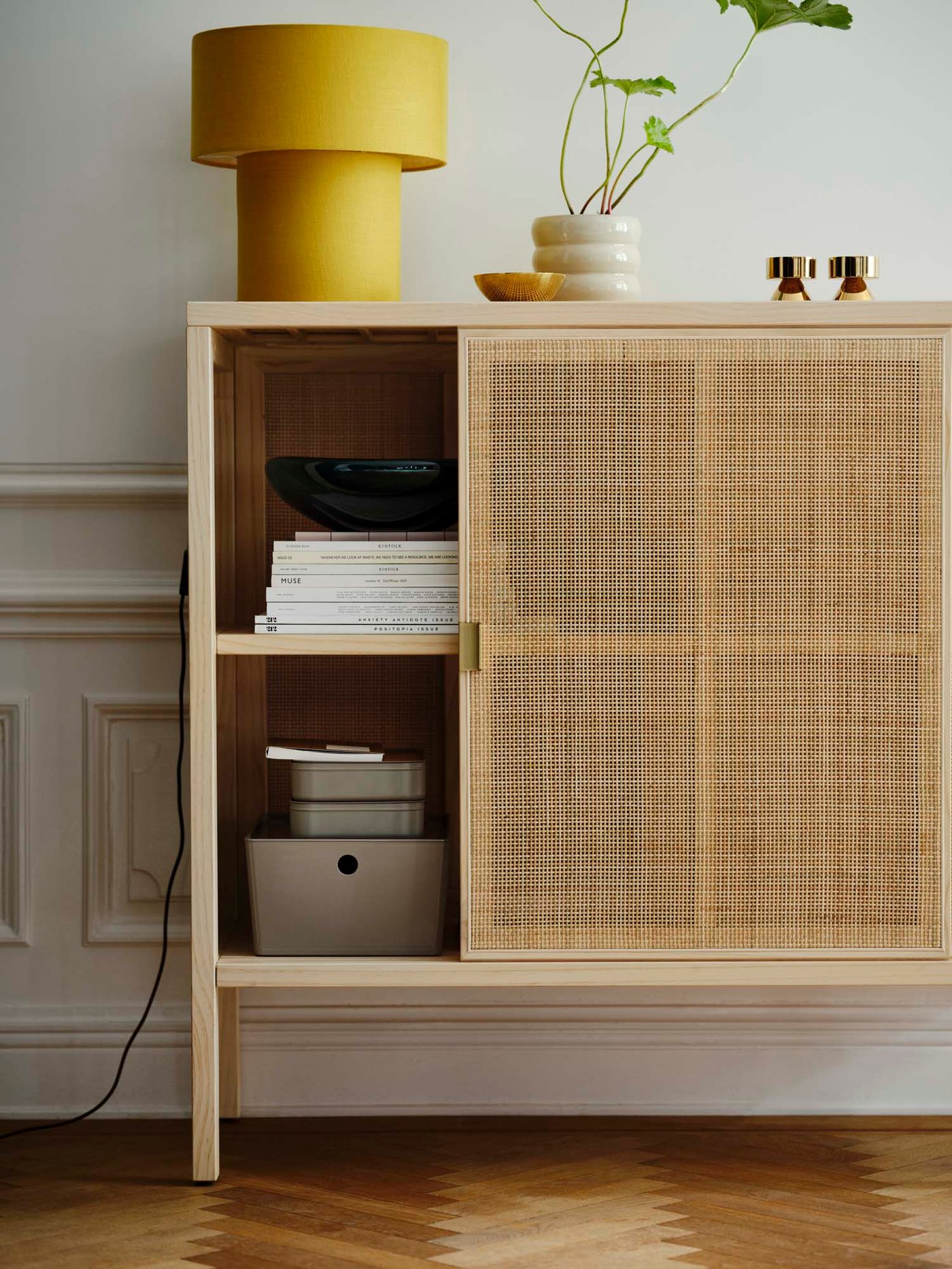 A rattan cabinet with an open sliding door with a yellow table lamp on top. 