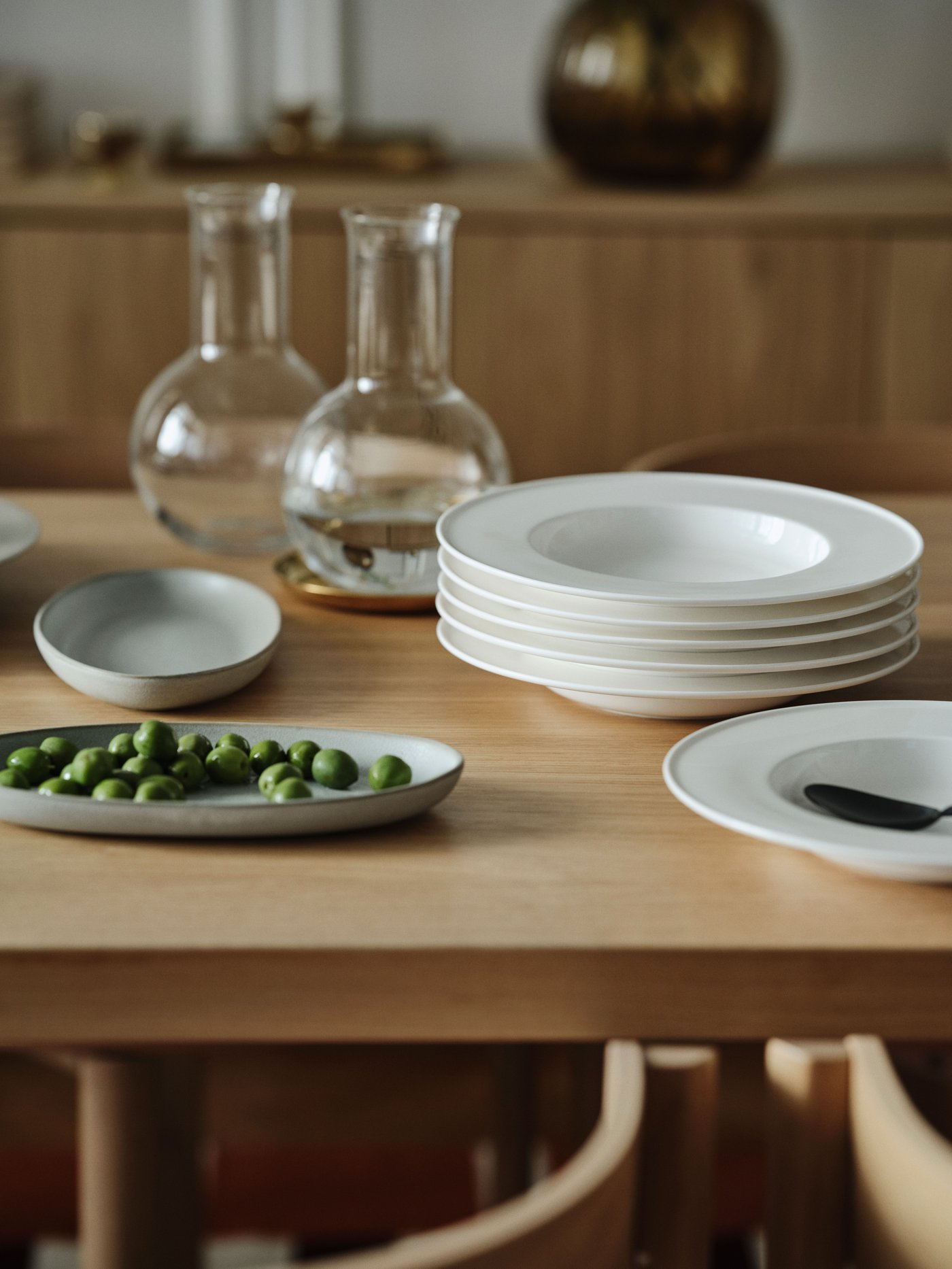 Stacked deep plates, serving bowls, and plates with greenery on a wooden table surface, glass carafes in the background.