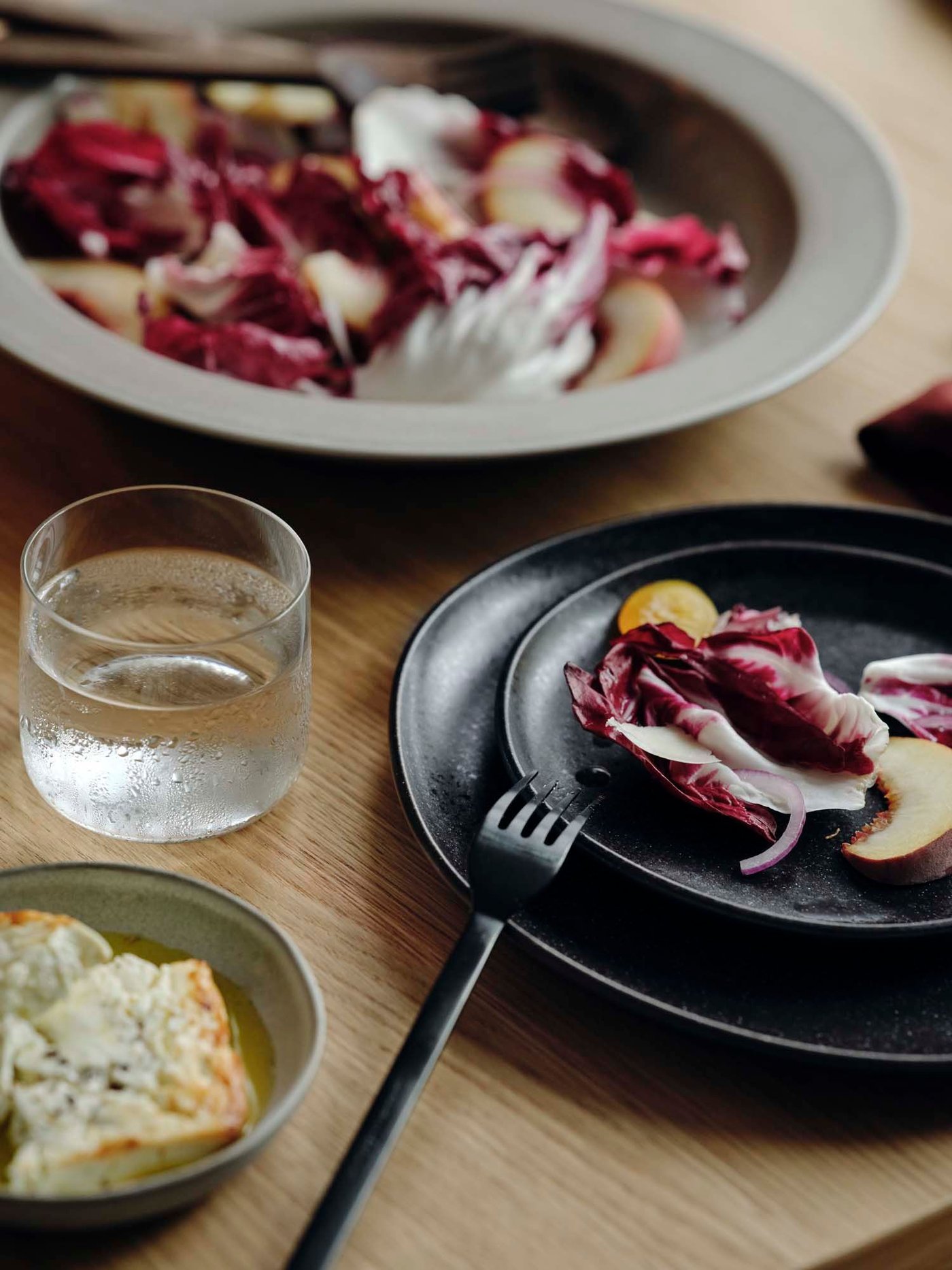 A close-up of dark and neutral coloured plates with food, next to forks and a glass.