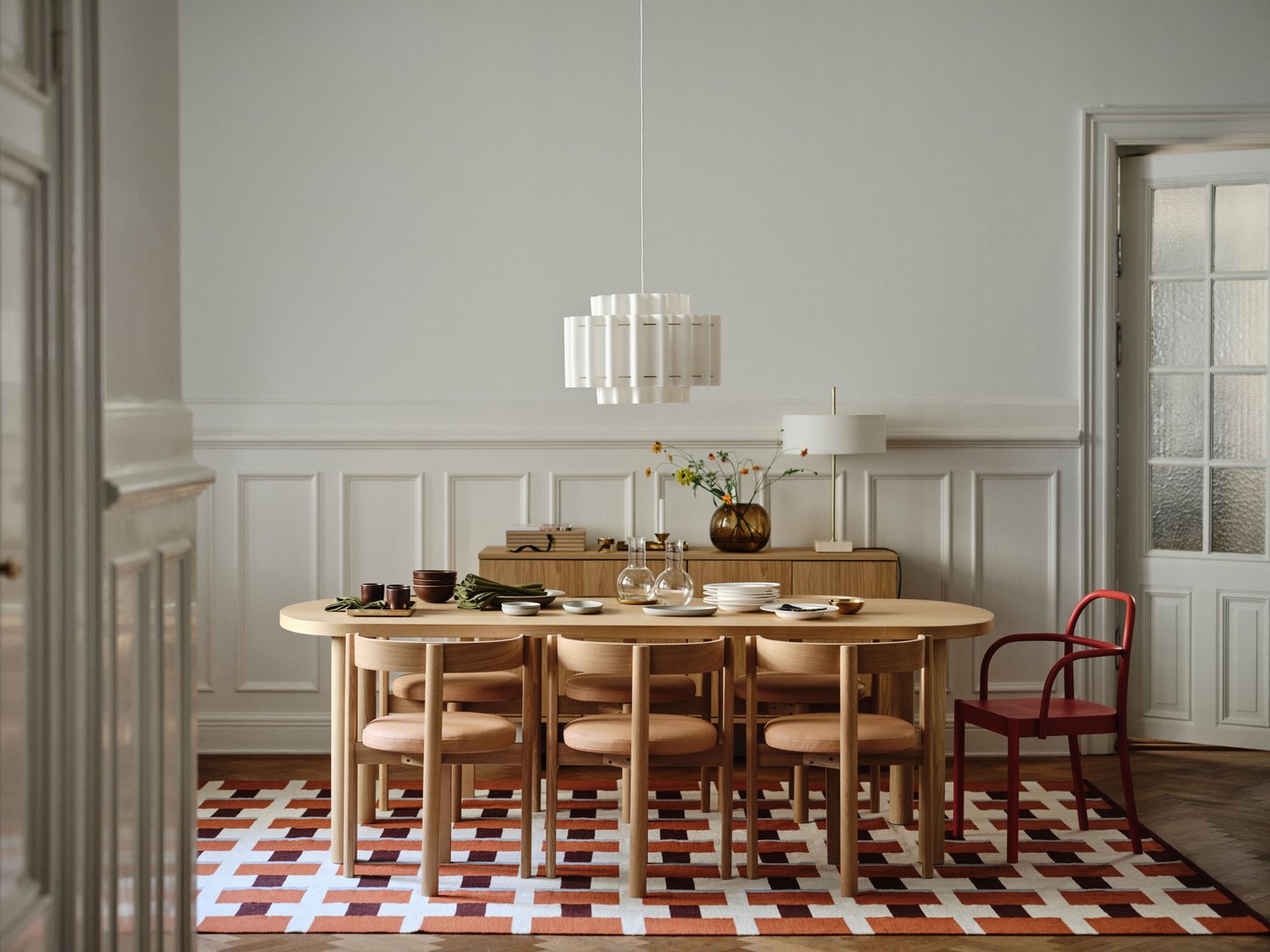 Long dining table on a burgundy rug in a classic Scandinavian apartment, with hanging lamp, gold accents, plates, leather-cushioned chairs, and a red accent chair.