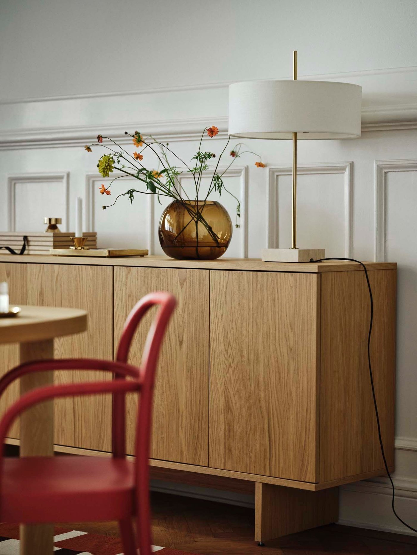 A wood storage unit with a vase and lamp, next to a red bent wood chair, from the STOCKHOLM collection.