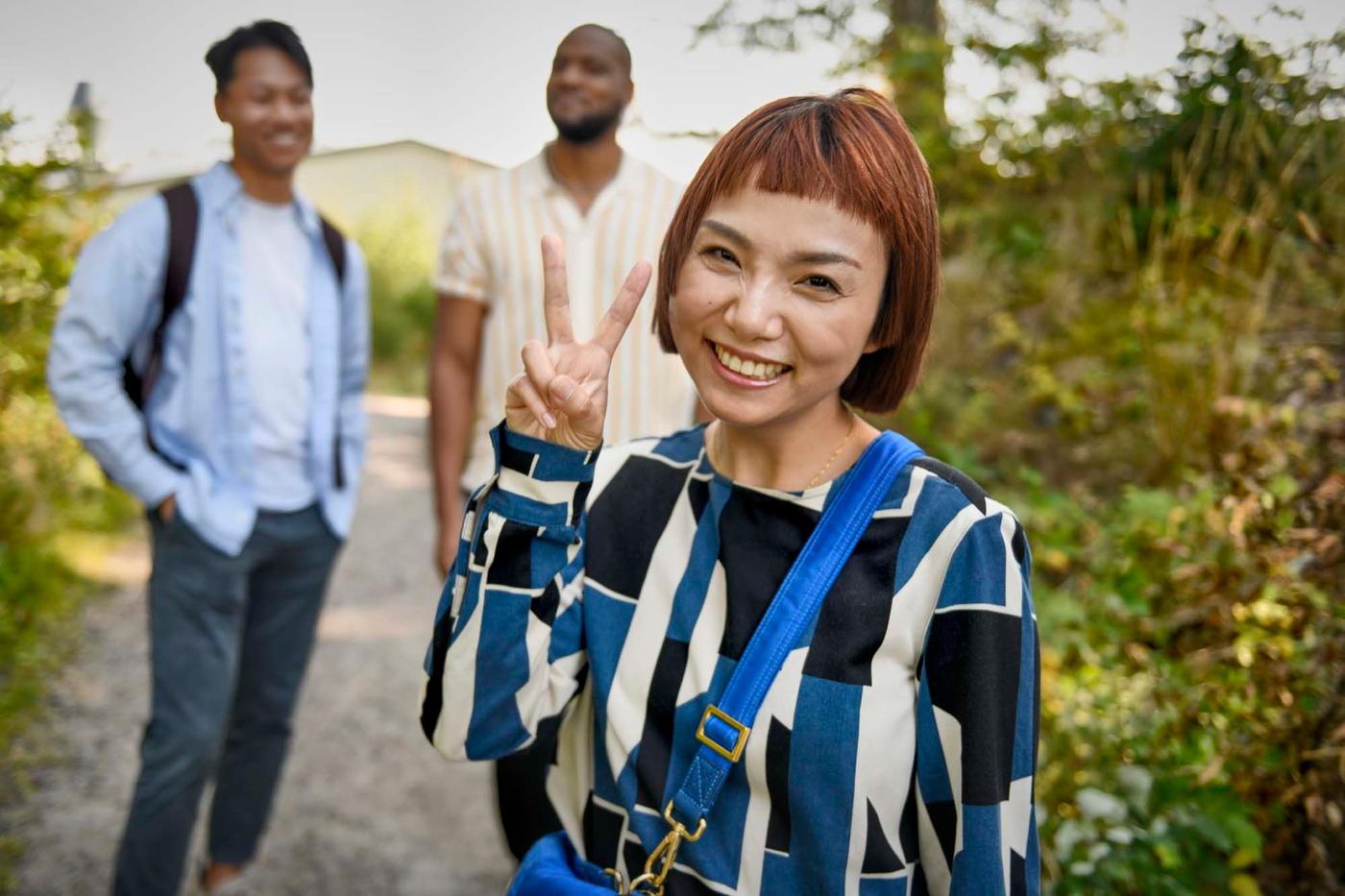 A person smiles at the camera and makes a victory sign, while two people stand in the background.