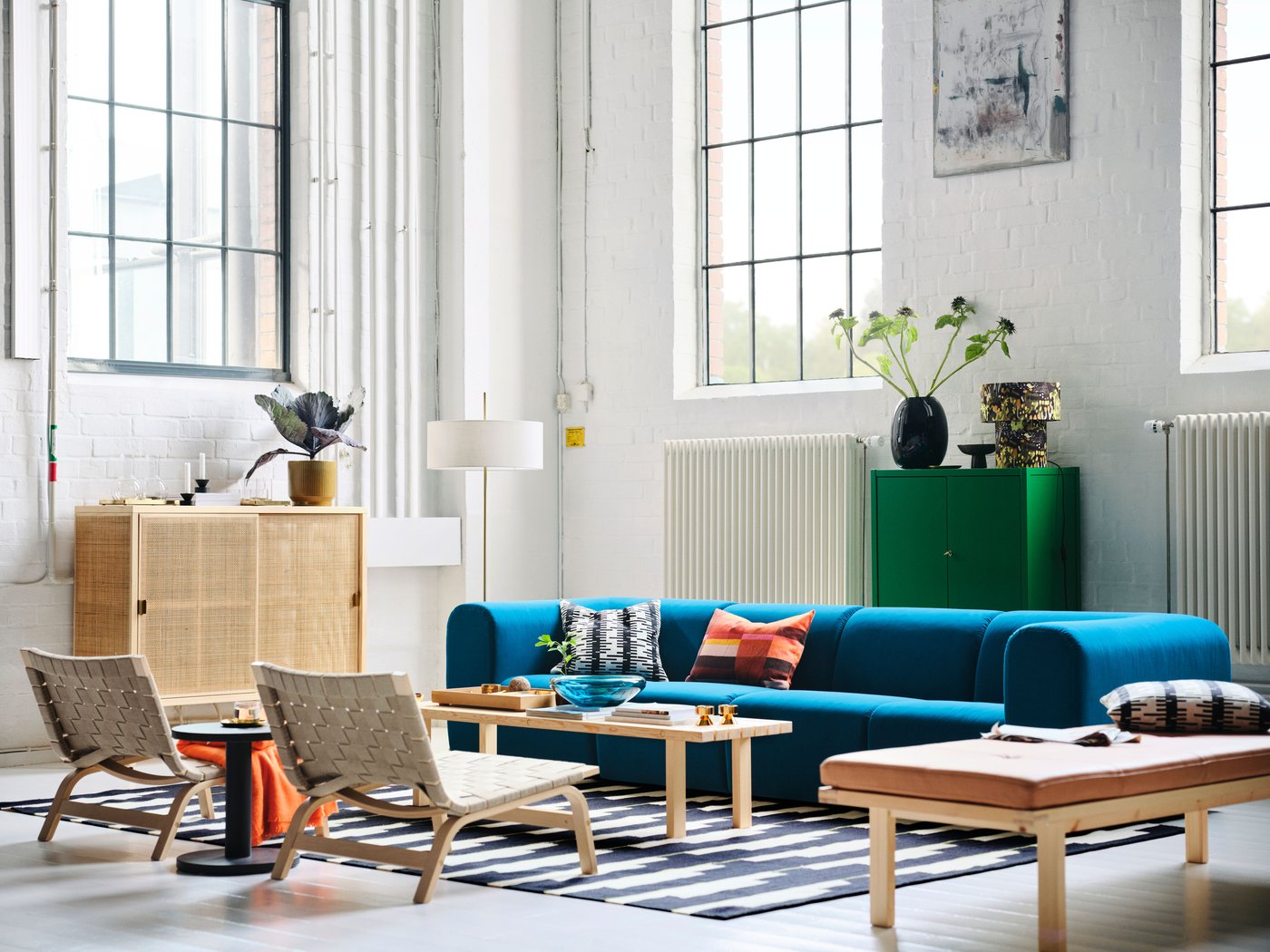 Blue-turquoise sofa in an industrial room with a daybed, saddle weave chairs, green oak cabinet, rattan cabinet, and large ceiling-high windows.