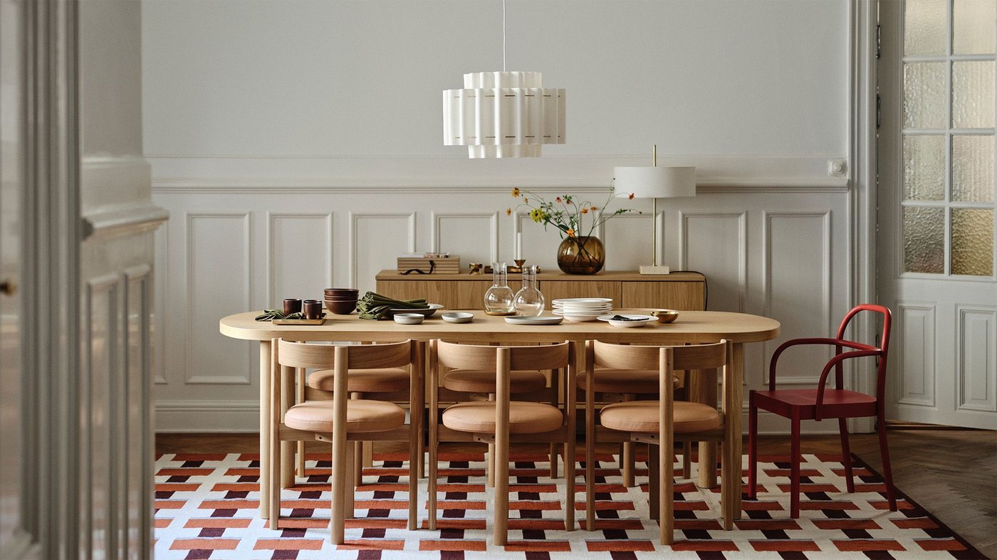 A classic style dining room with white panelled walls. A white lamp hangs over a wood table and chairs and a colourful patterned rug, all from the STOCKHOLM 2025 collection.