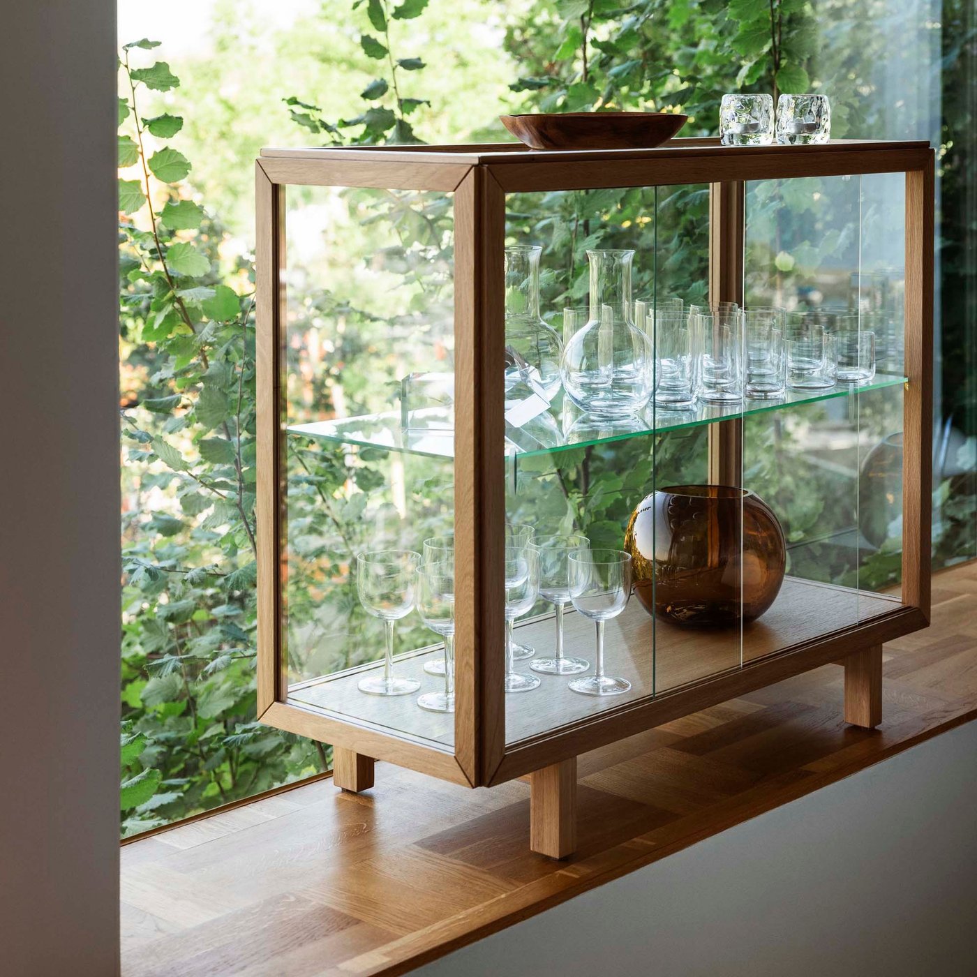 An oak veneer STOCKHOLM 2025 glass-door cabinet holding glassware, in front of a large window with a leafy outlook.