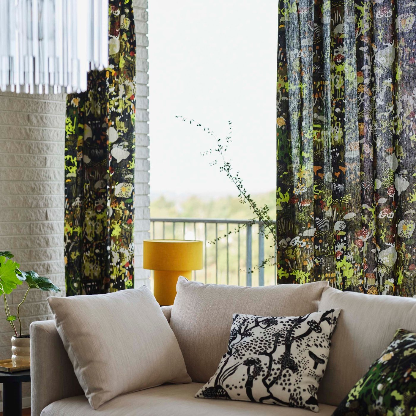 A light coloured sofa with cushions and a yellow lamp, in a room against a window and floral curtain background.