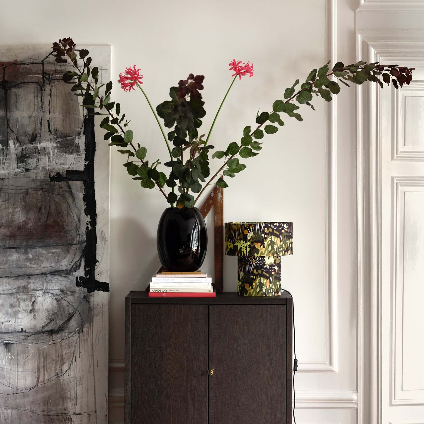 A large black STOCKHOLM vase filled with long stemmed foliage and flowers sits on a dark brown storage unit in a living room..