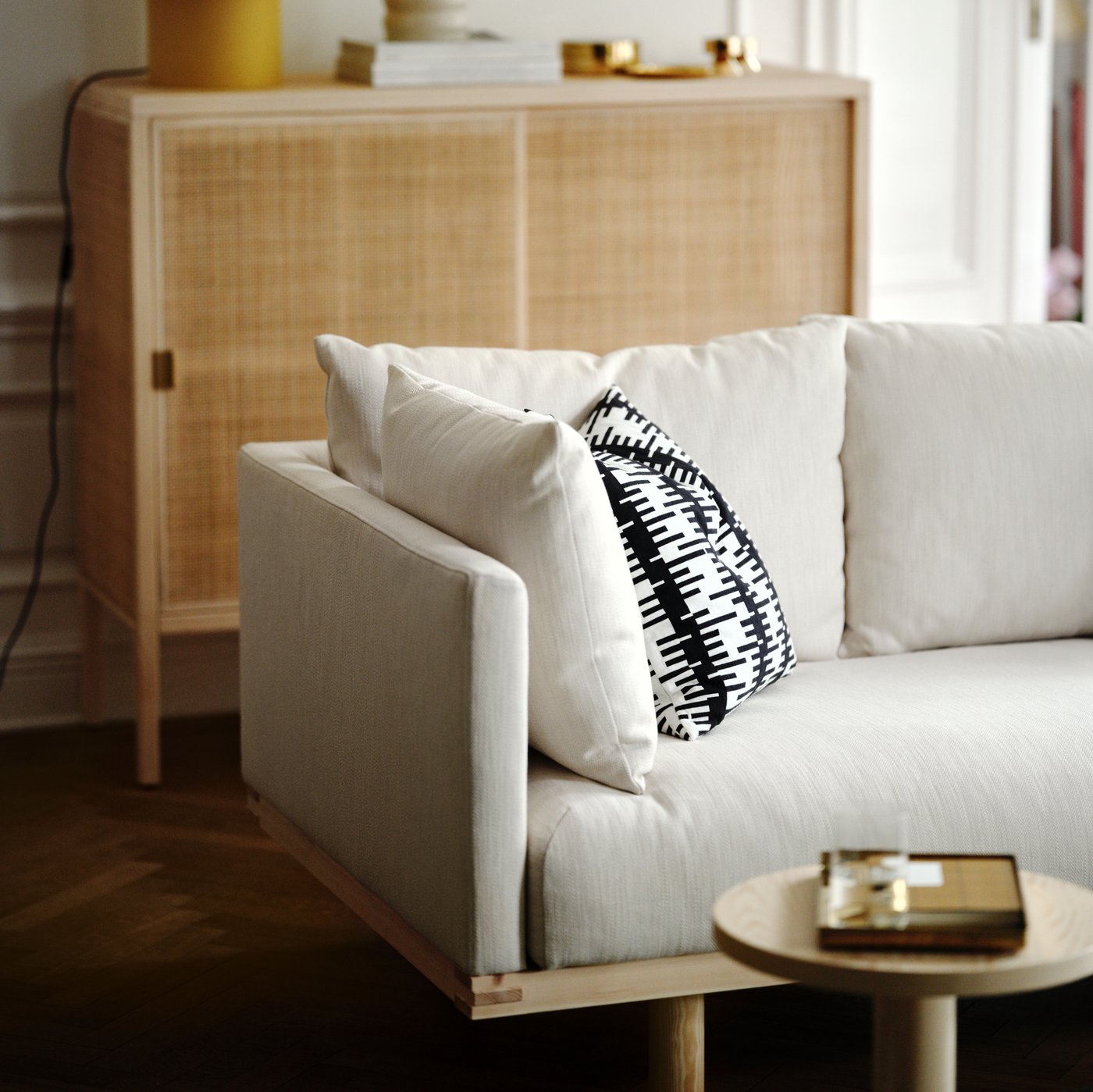 Close-up of beige wool sofa with cushions, table in front and rattan cabinet in the background.