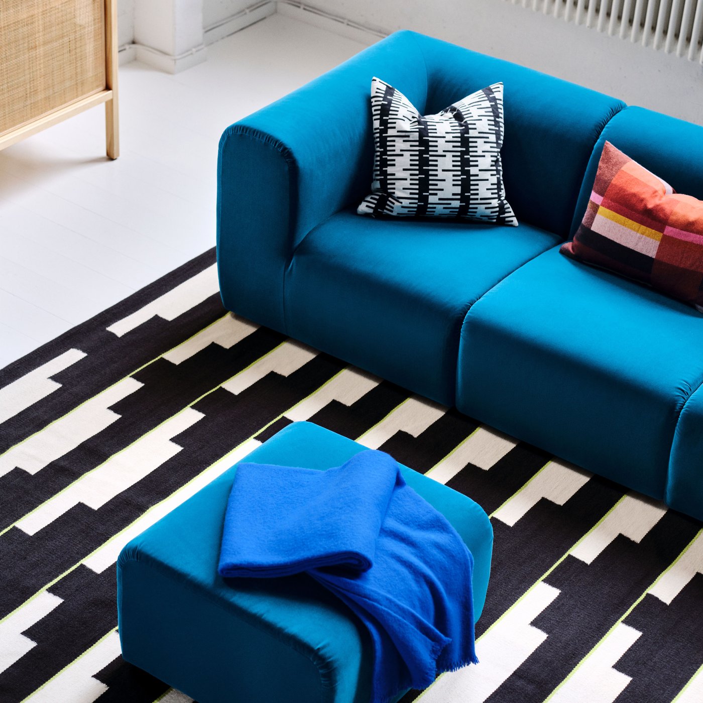 Top view of turquoise sofa with two checked pillows on black and white rug, paired with matching pouffe.