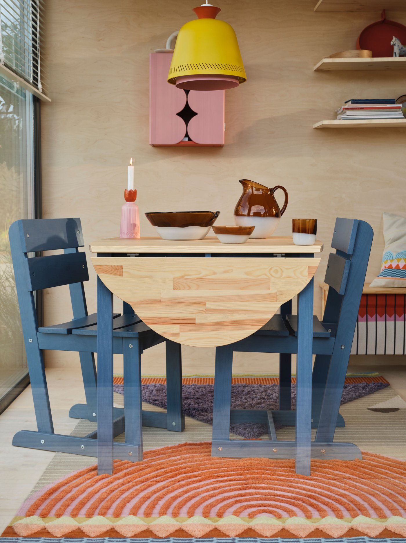 A table and two blue chairs stand in a wood and glass roomset, with a yellow lamp hanging above.