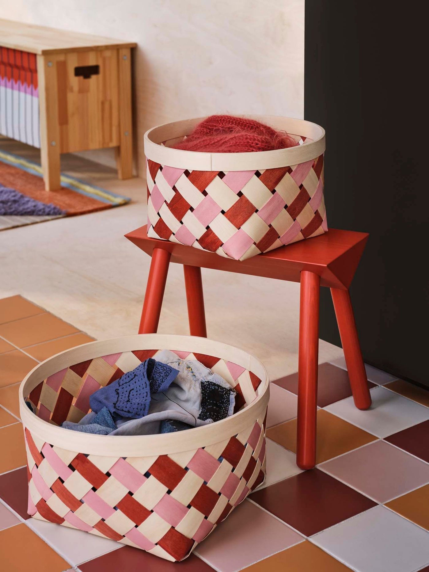 A small red, pink and natural chequered basket sits on a stool next to a similar, larger basket. They are filled with textiles.