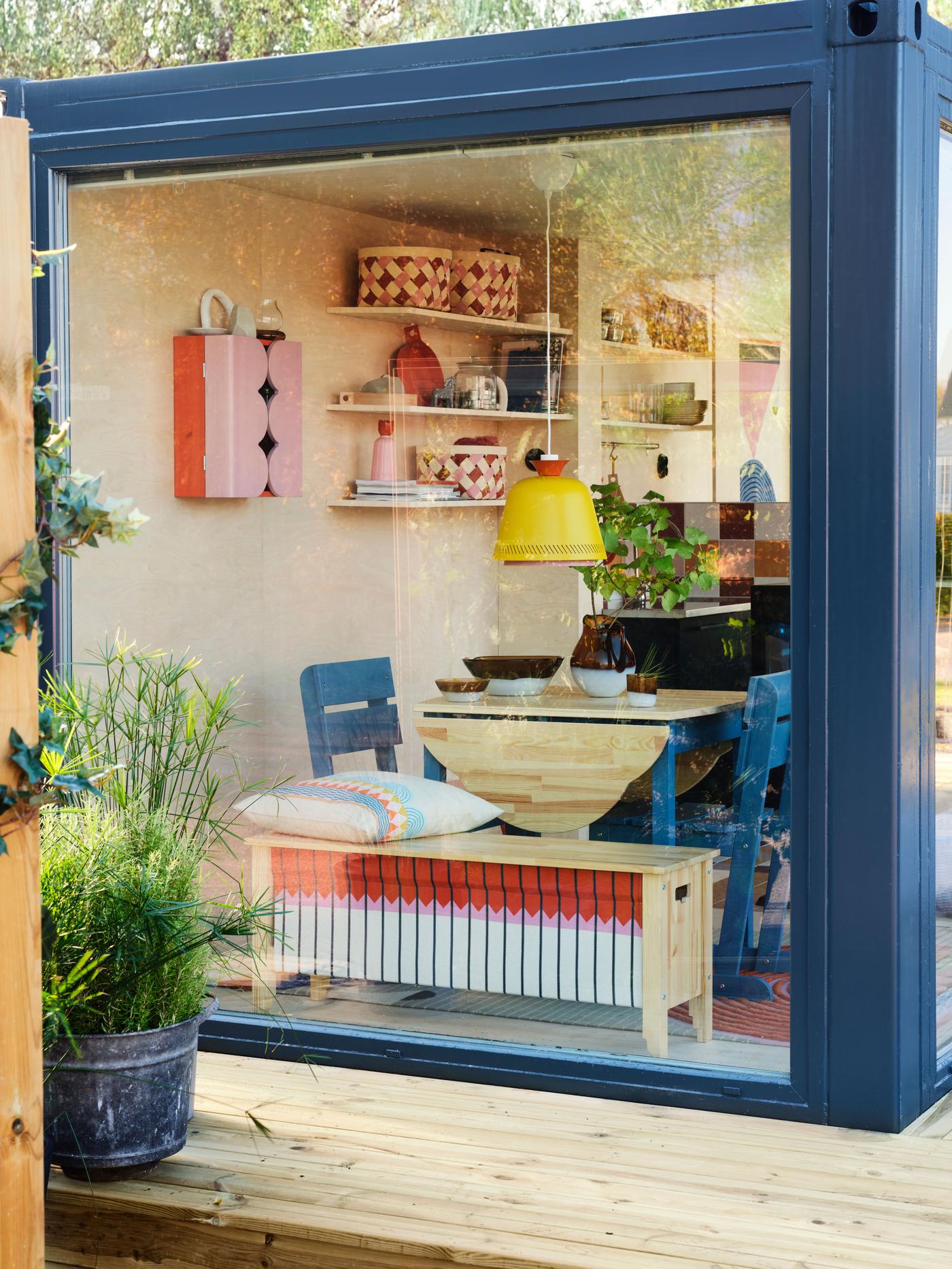 Chairs, tables and other items from the OMMJÄNGE collection, seen through a blue-framed window of a cabin-like container house. 