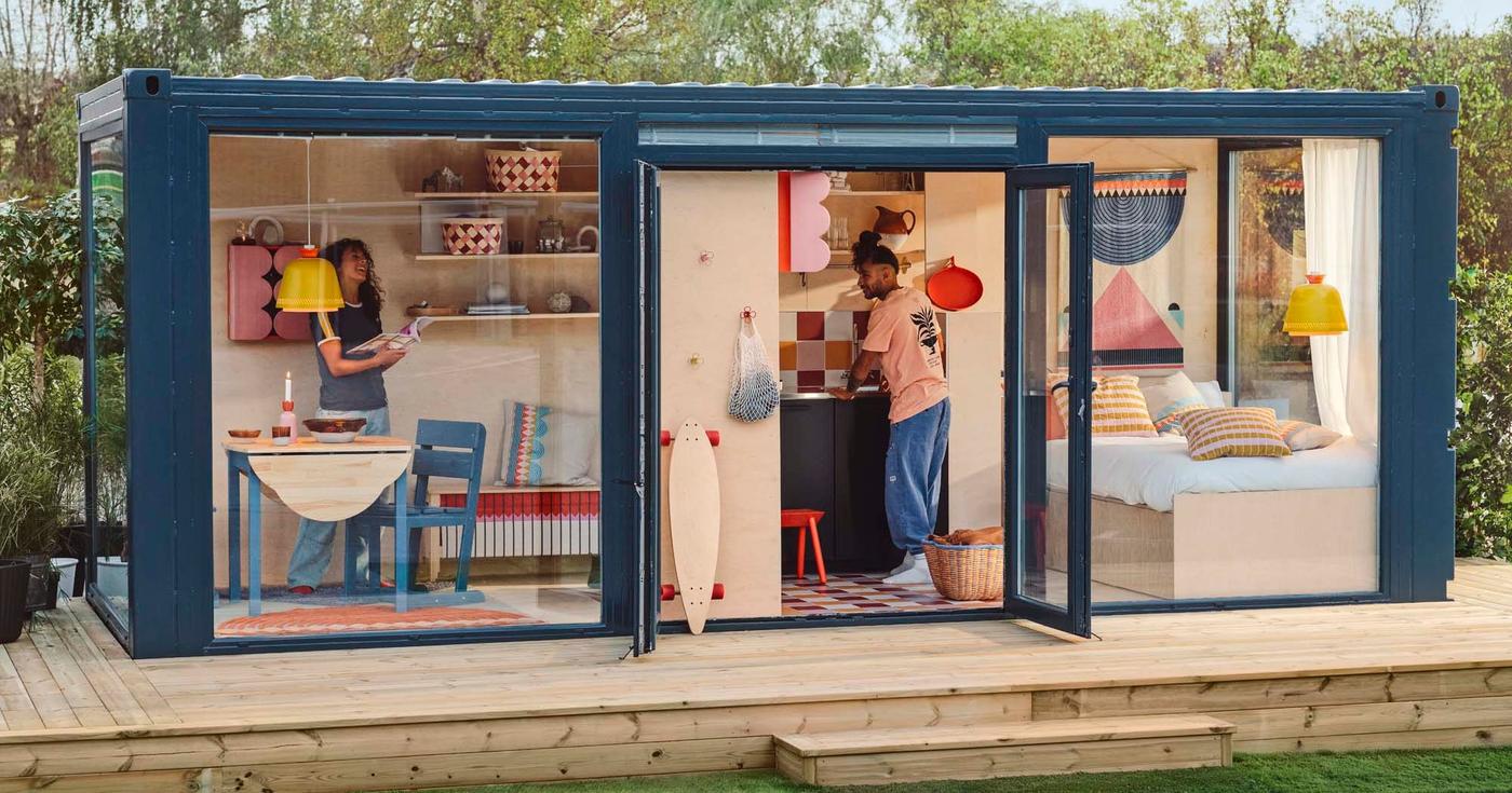 Two people stand in a colourful roomset inside a blue framed wood and glass cube. The cube is outdoors and stands on a wood deck.