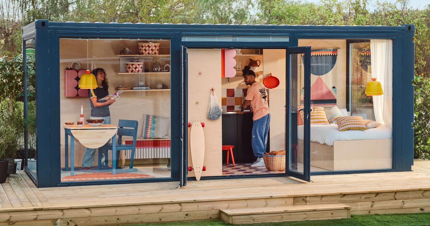 Two people stand in a colourful roomset inside a blue framed wood and glass cube. The cube is outdoors and stands on a wood deck.