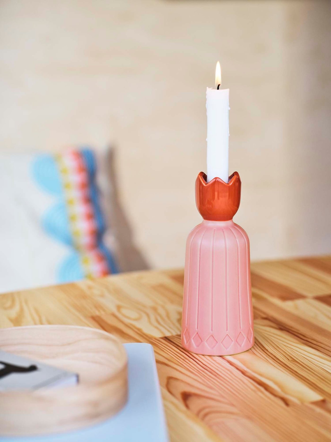 A pink and orange OMMJÄNGE candlestick with an illuminated candle stands on a wood table with a cushion in the background.