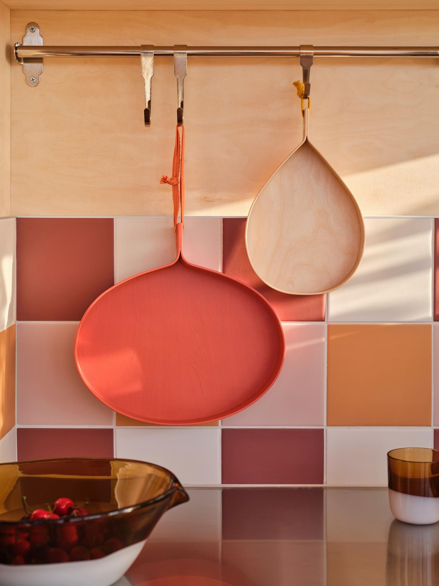 A large red/orange and a smaller wood tray hang from a rail against a wood and tiled wall.