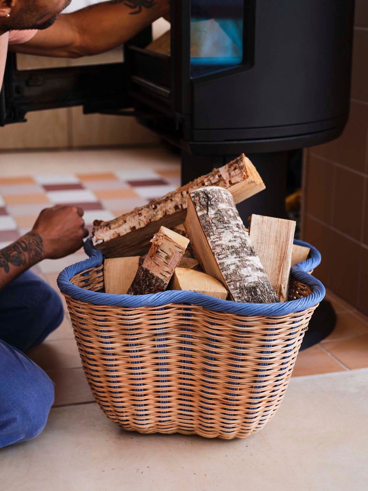 A blue and wicker basket filled with wood stands on the floor.