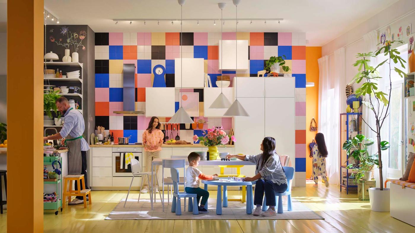 A sunny and colourful kitchen in orange, blue and white tones with a parent and child sitting at a children's table and another child standing in the kitchen.