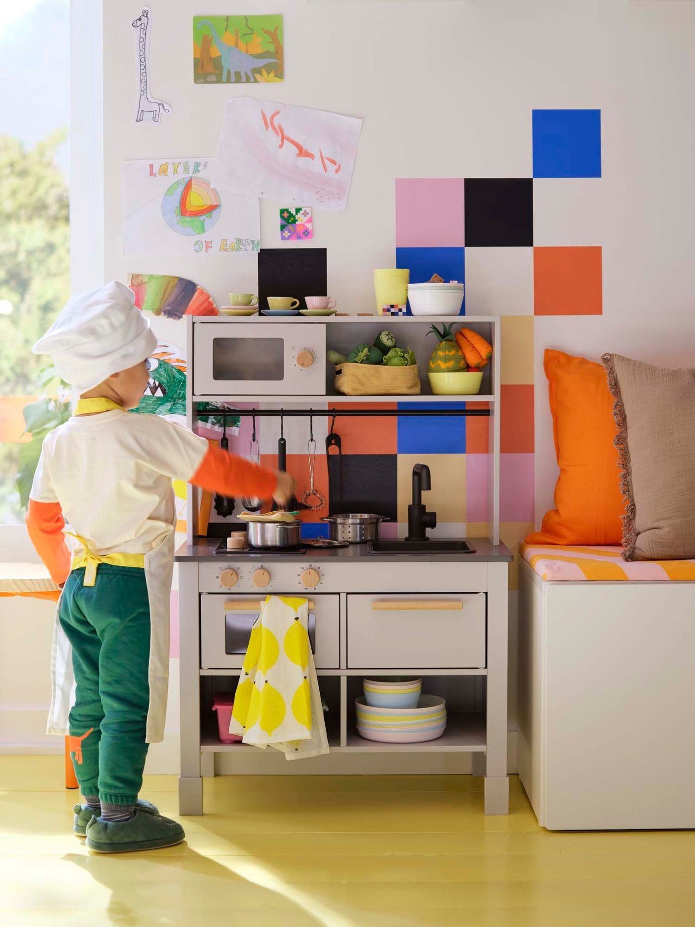 A young child wearing an apron and chefs hat stands beside a white toy kitchen in a colourful room.