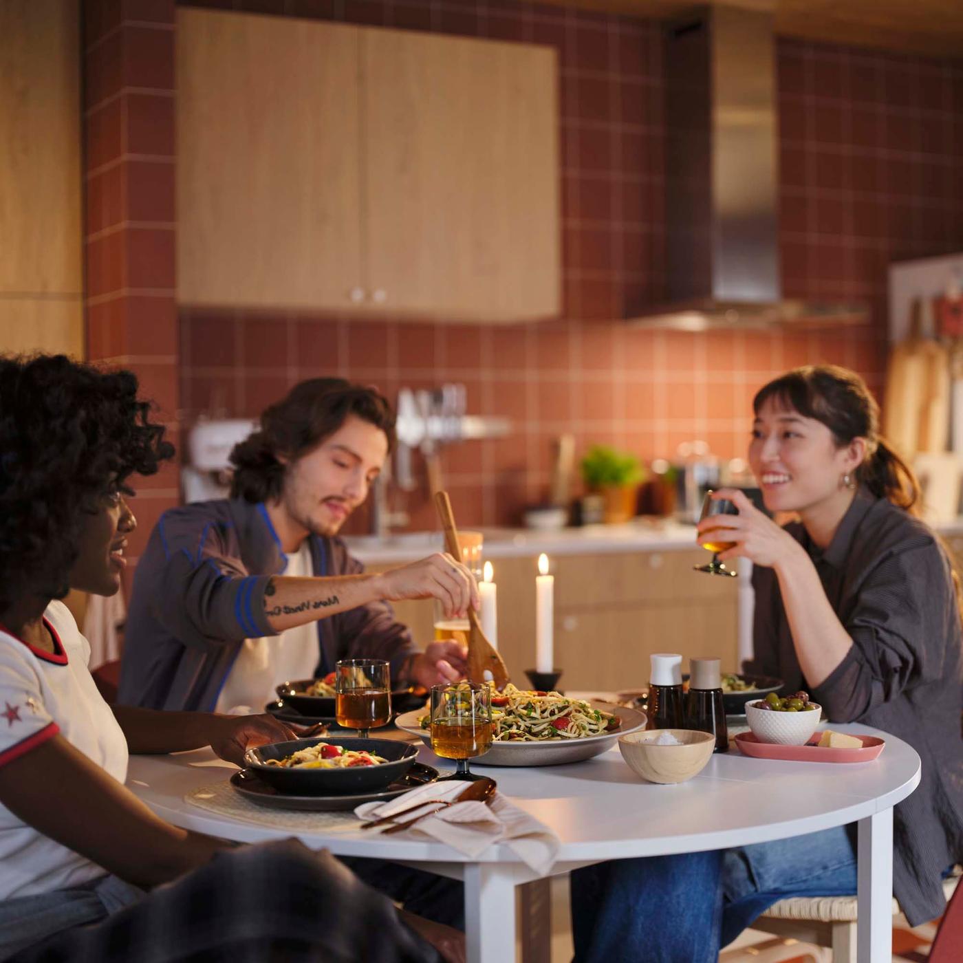 Three people enjoying pasta together in an oak-effect KNOXHULT kitchen with burnt orange tiles.