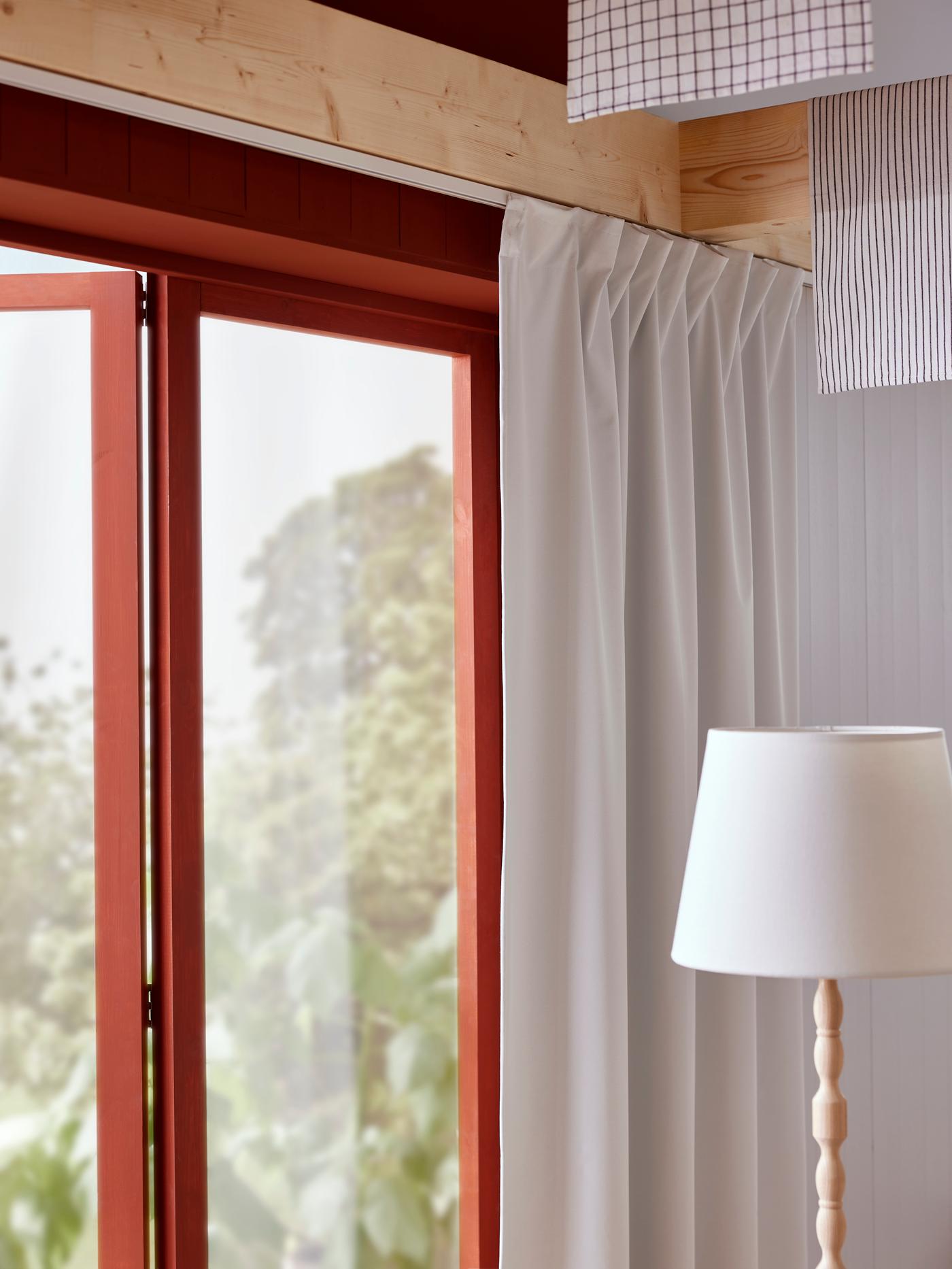 Close up of an open, red-framed window with white curtains and a white-shaded floor lamp nearby.