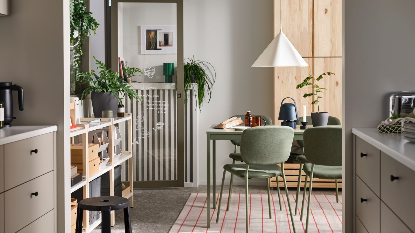 A view from the kitchen into a neutral coloured dining room with plenty of potted plants, green furniture an wood cupboards.