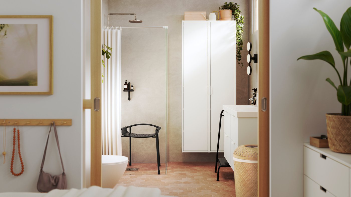 View of a bathroom with a curtained shower enclosure and chair and white storage units, seen from the bedroom. There is a plant on top of the storage unit.