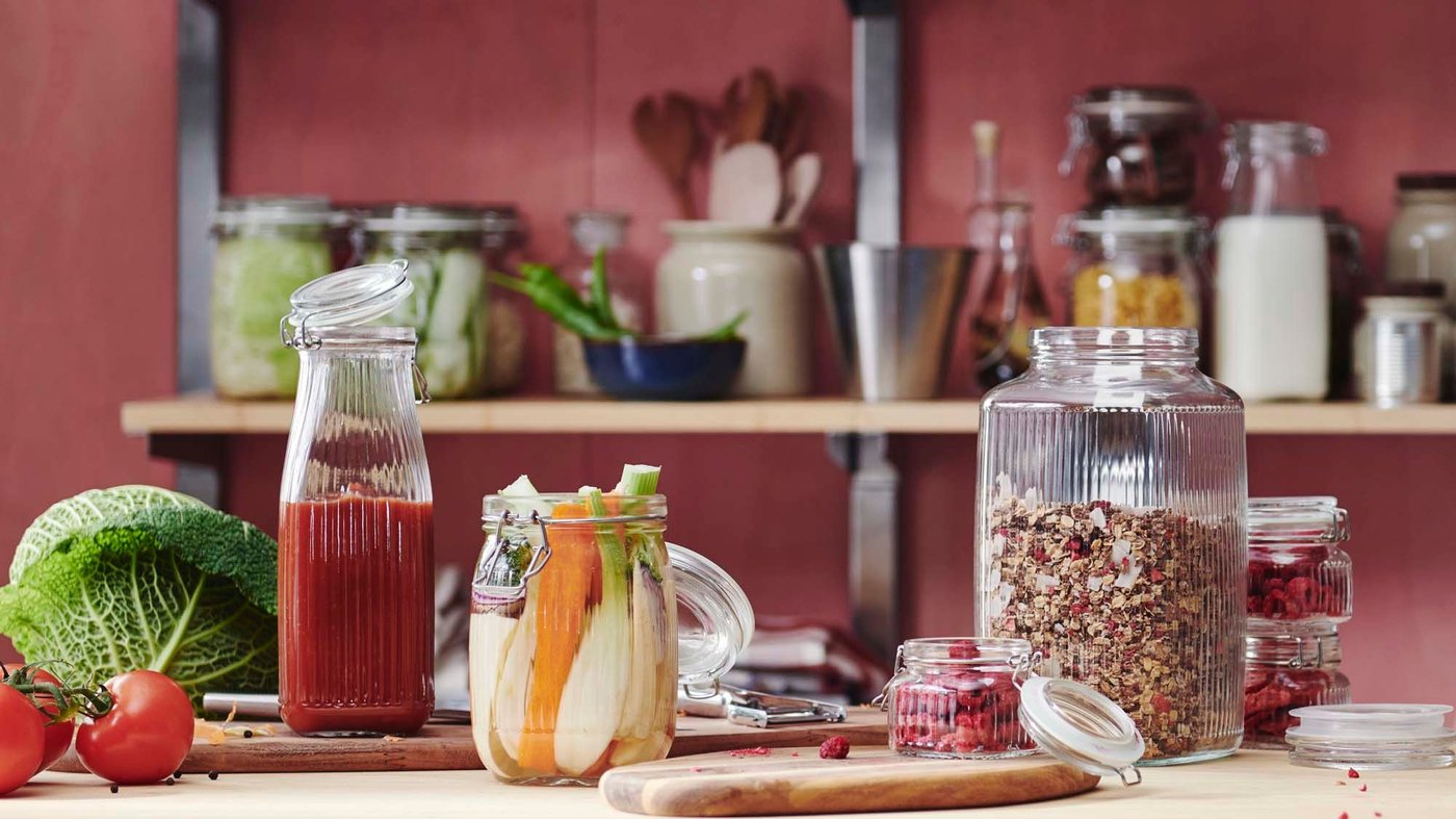 A bottle filled with blended tomato, and jars filled with vegetables, raspberries and granola sit on a wooden table, with jars in the background.