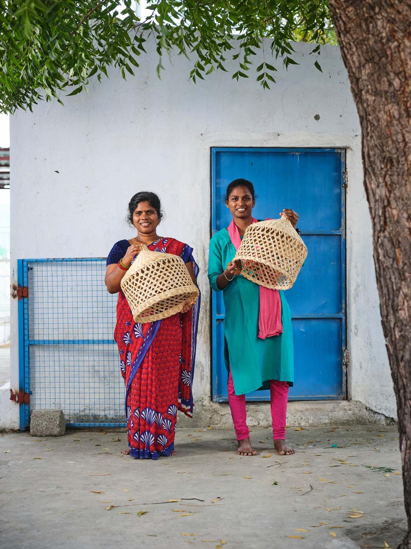 Two women stand in front of a white building with a blue door. They are each holding handmade rattan lampshades.